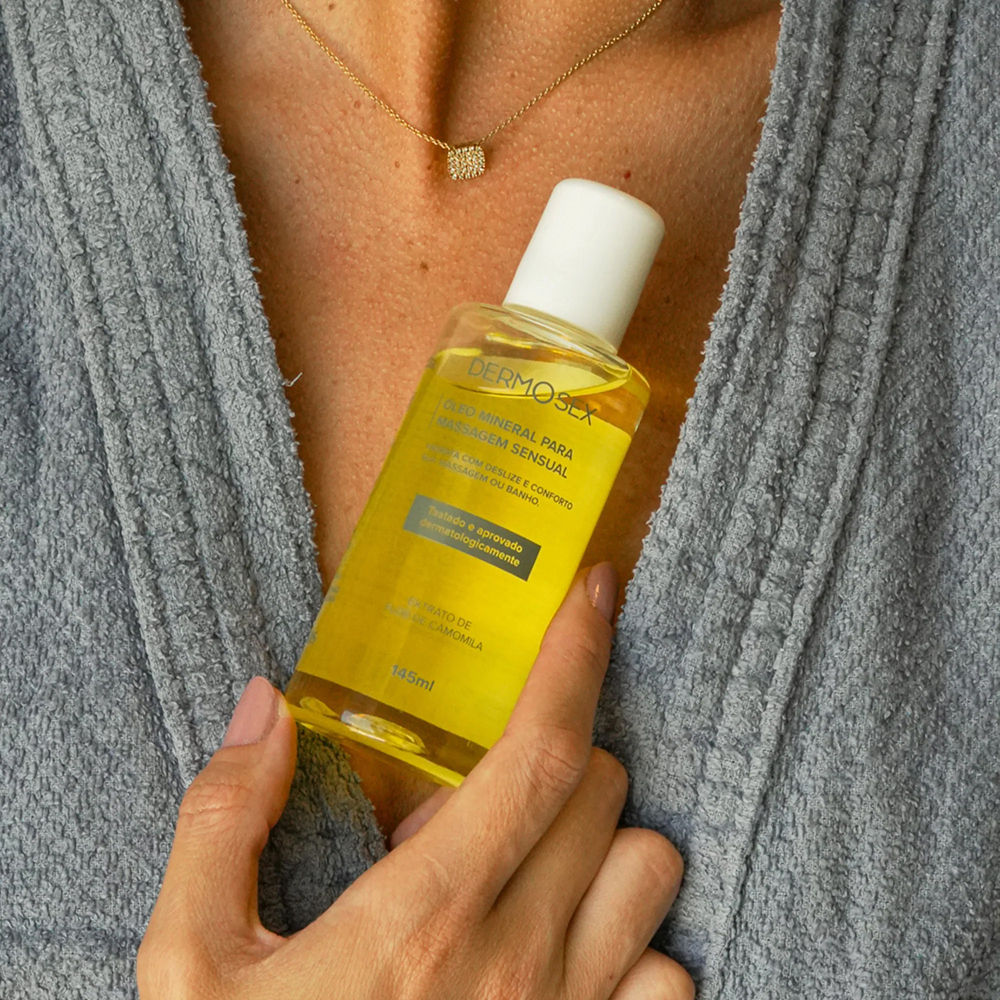 �leo Mineral para Massagem Sensual com Extrato de Flor de Camomila 145ml, sendo segurado por uma m�o, com um robe ao fundo