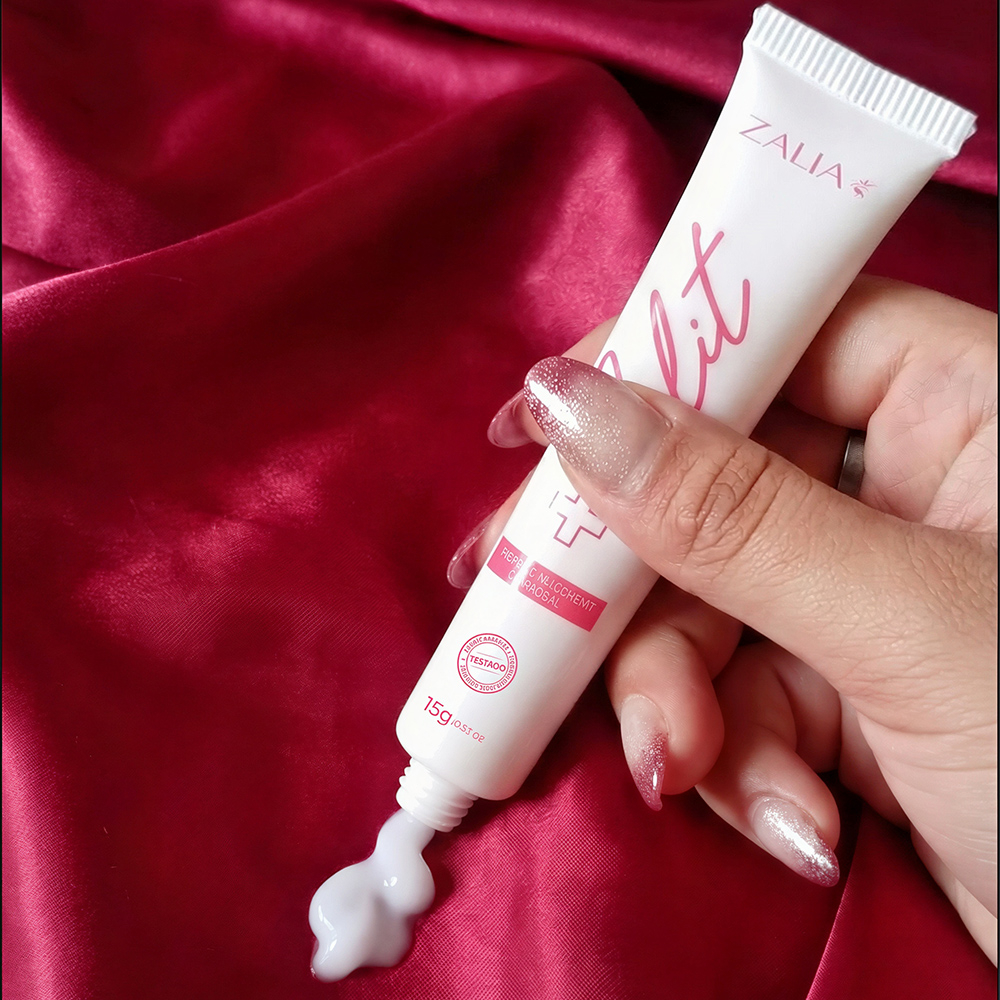Close-up de m�o feminina apertando o Gel Excitante Feminino Clit da Zalia Cosm�tico, mostrando a textura do gel branco em um fundo de tecido vermelho